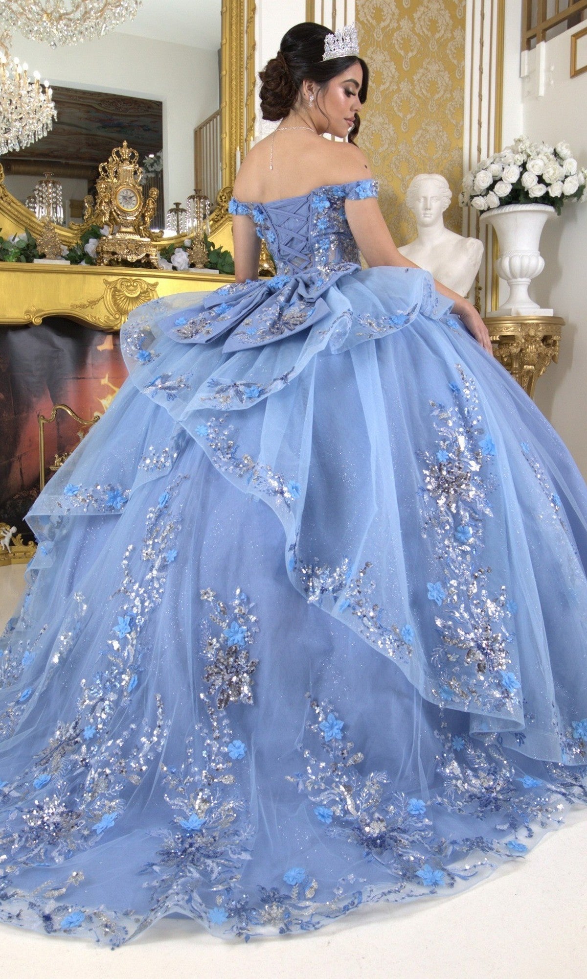 A woman wearing the Quinceanera Ball Gown 8125J with floral embellishments and a tiara stands indoors, facing away from the camera in an ornate room featuring gold accents and a fireplace.
